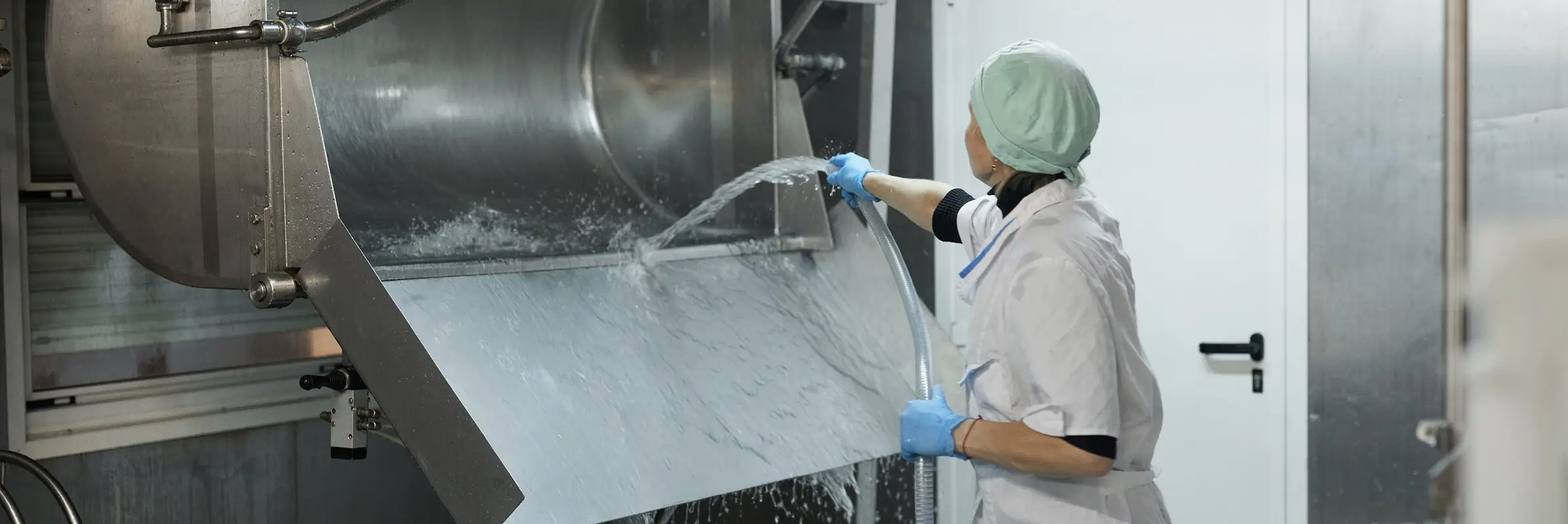 Food processing worker in white uniform, hairnet, and blue gloves hosing down industrial mixing equipment in a commercial facility