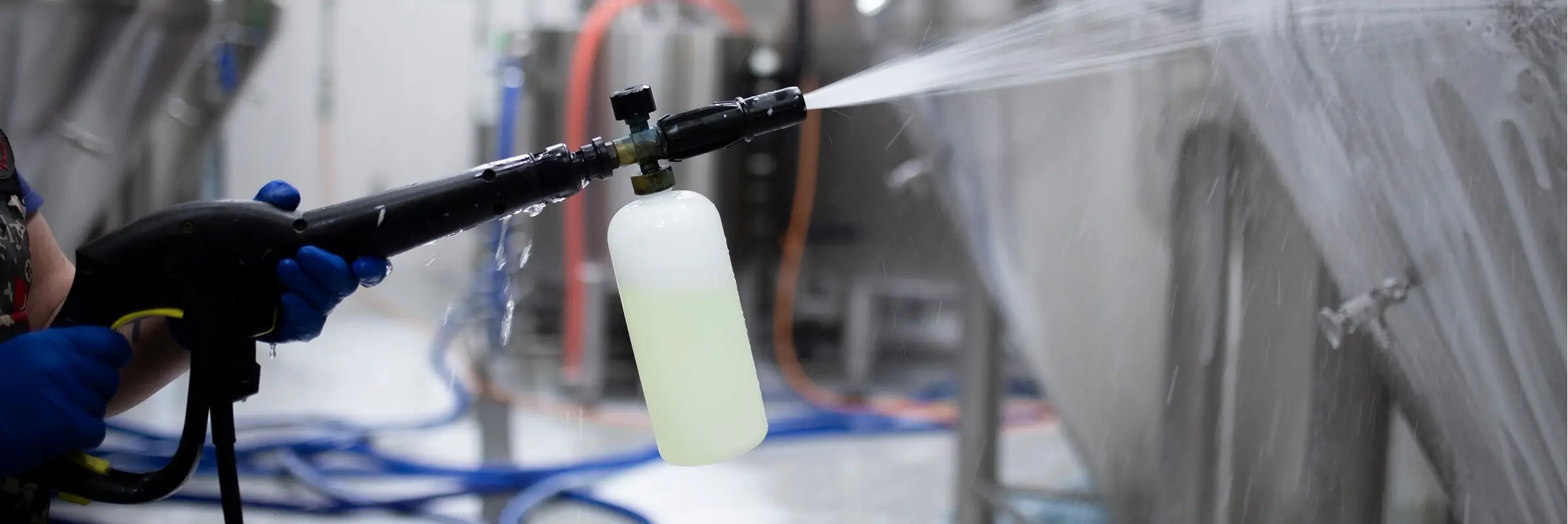Close-up of a foam cannon spray gun with detergent bottle applying cleaning solution to stainless steel industrial tanks