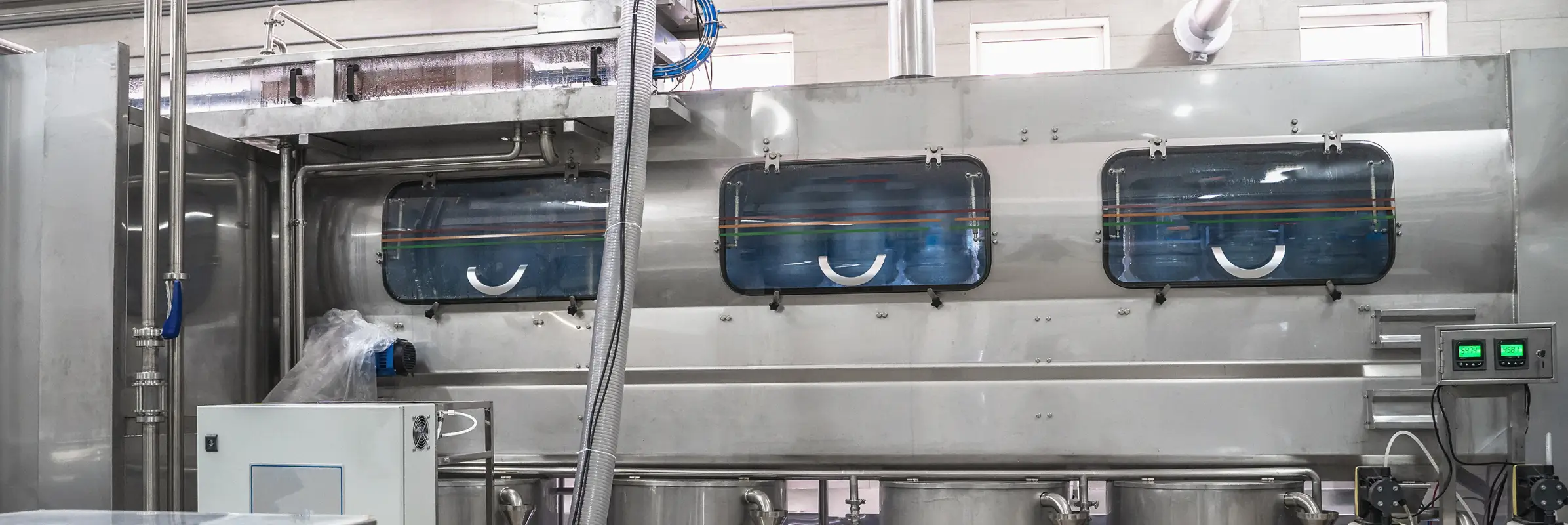 Large stainless steel industrial processing machine with three observation windows, connected hoses, and digital control panels in a factory setting